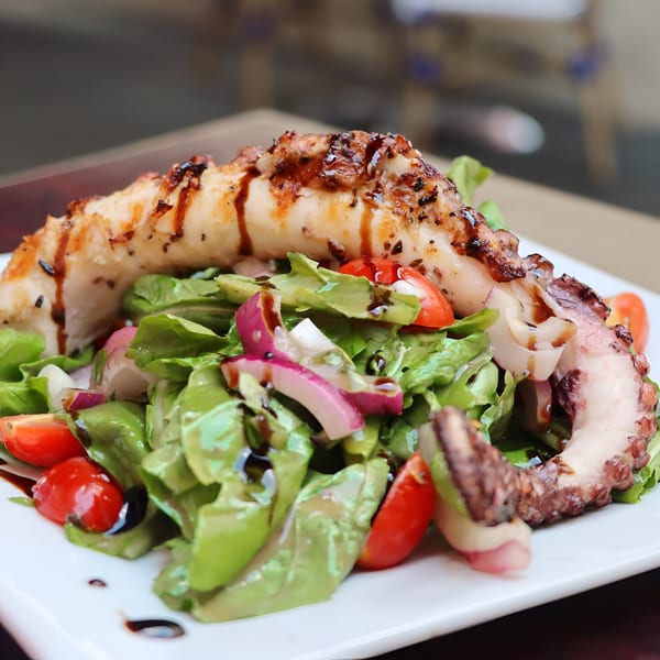 Grilled octopus with a fresh green salad, cherry tomatoes, and red onions, drizzled with balsamic glaze.