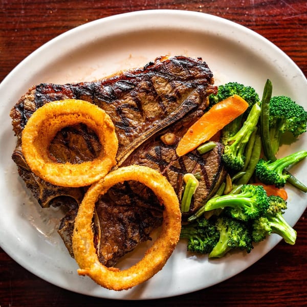 A grilled T-bone steak with two onion rings, broccoli, green beans, and carrots on a white plate.