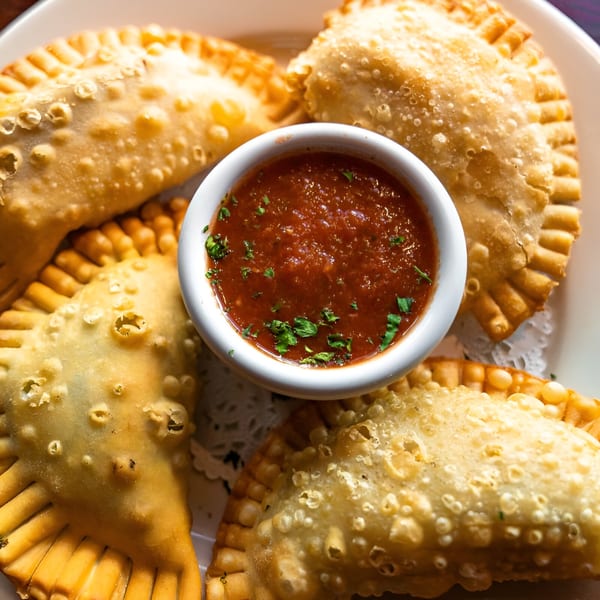 Golden-brown empanadas with crimped edges arranged around a white bowl of red dipping sauce.