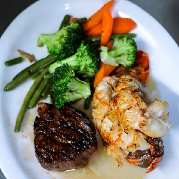 A surf and turf dish with steak, lobster tail, broccoli, green beans, and carrots.